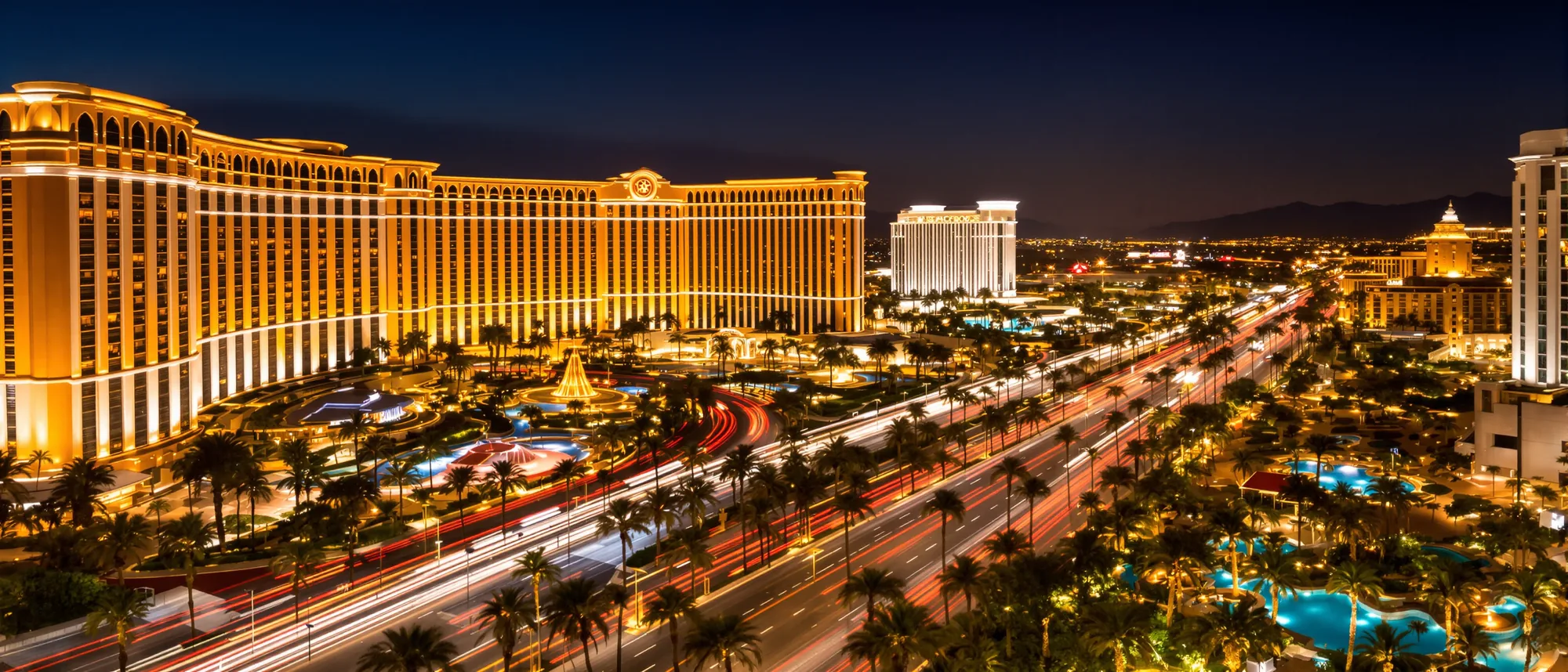 A resort boulevard at night seen from above, glowing hotel towers, palm trees, pools and streaks of traffic light