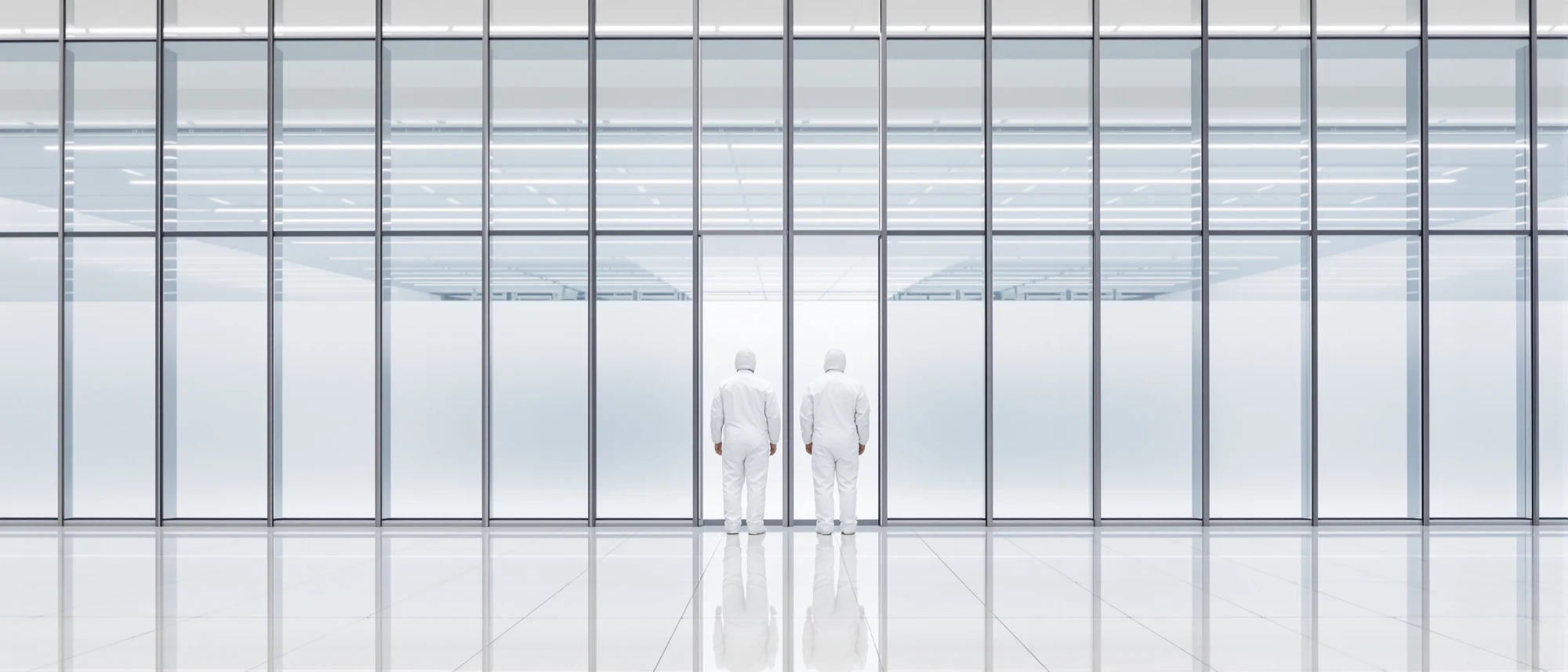 Two technicians in white clean room suits standing before a vast glass wall into a sealed server hall