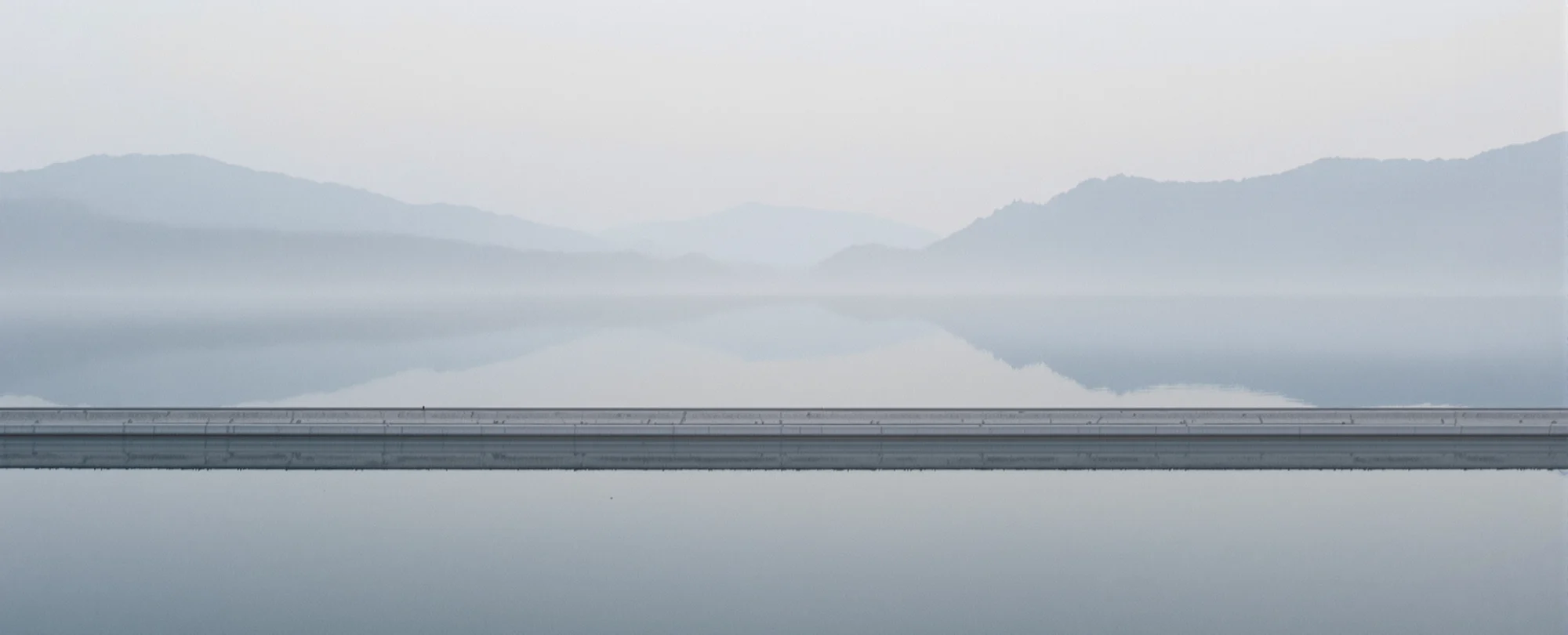 A still mountain reservoir in morning mist, held by a long concrete dam crest