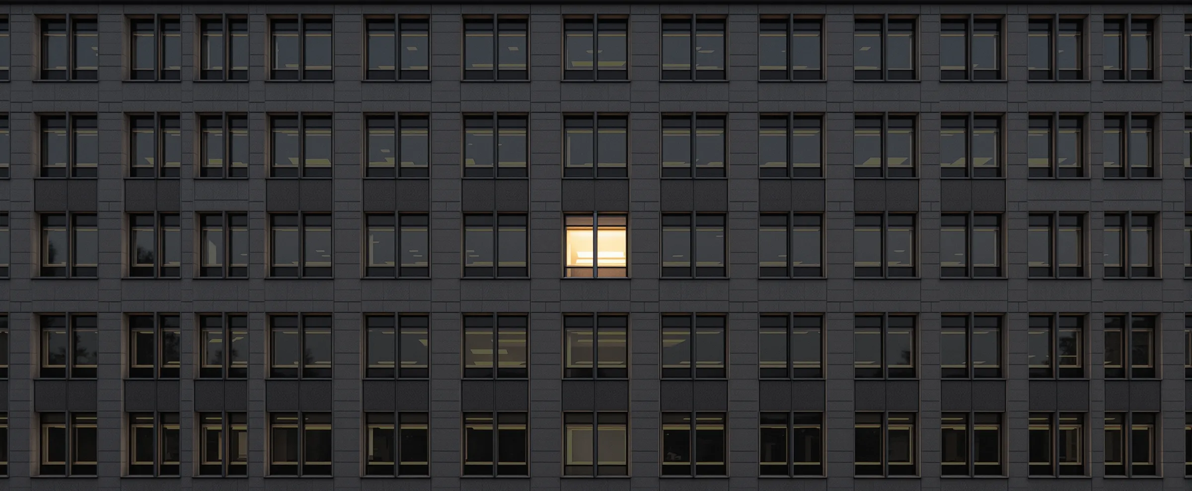 A straight-on view of an office building facade after sunset, a grid of dark windows with a single lit office in the center