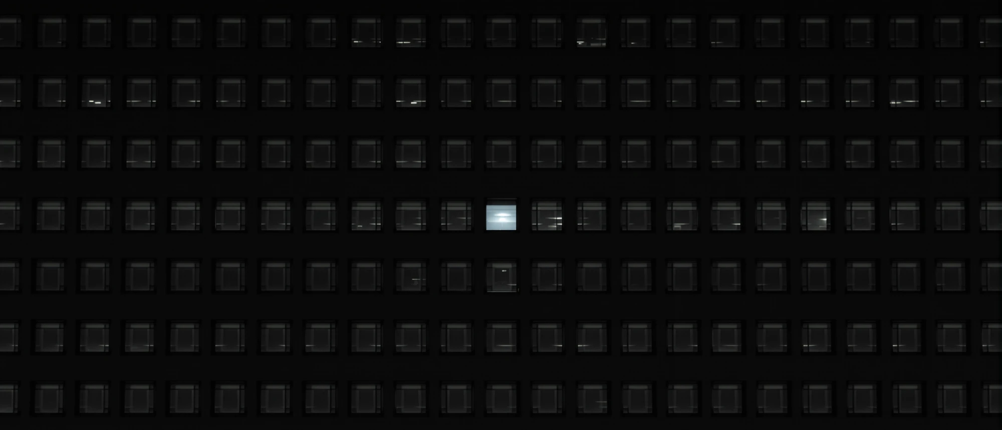 The facade of a large office building at night, a grid of dark windows with a single window lit white